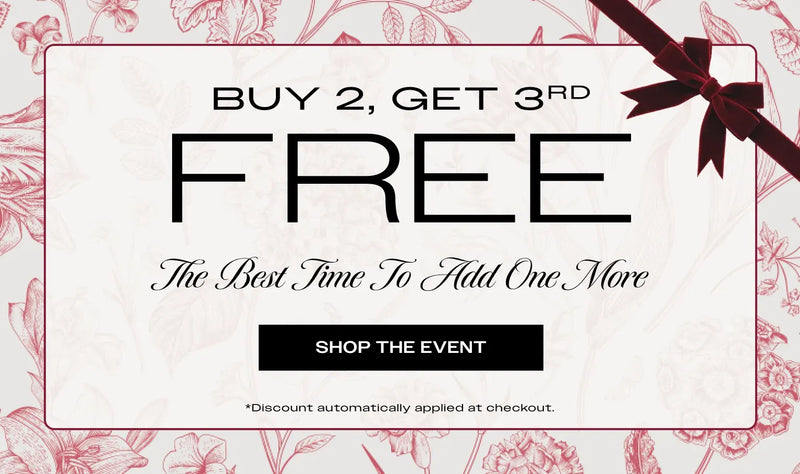 Promotional graphic banner with the text 'BUY 2, GET 3RD FREE' , with a subtitle reading 'The Best Time To Add One More,' with a 'SHOP THE EVENT' button , and fine print at the bottom reads 'Discount automatically applied at checkout' . The design has a white central panel with a thin border, red floral line art around the edges, and a dark red ribbon bow in the top-right corner.