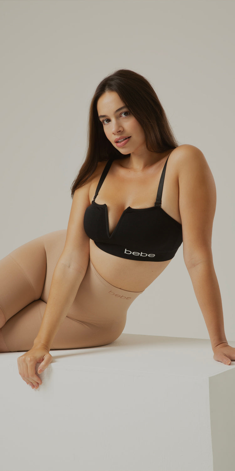 A woman wearing a black bebe bra and beige tights poses on a white platform against a light gray studio background.